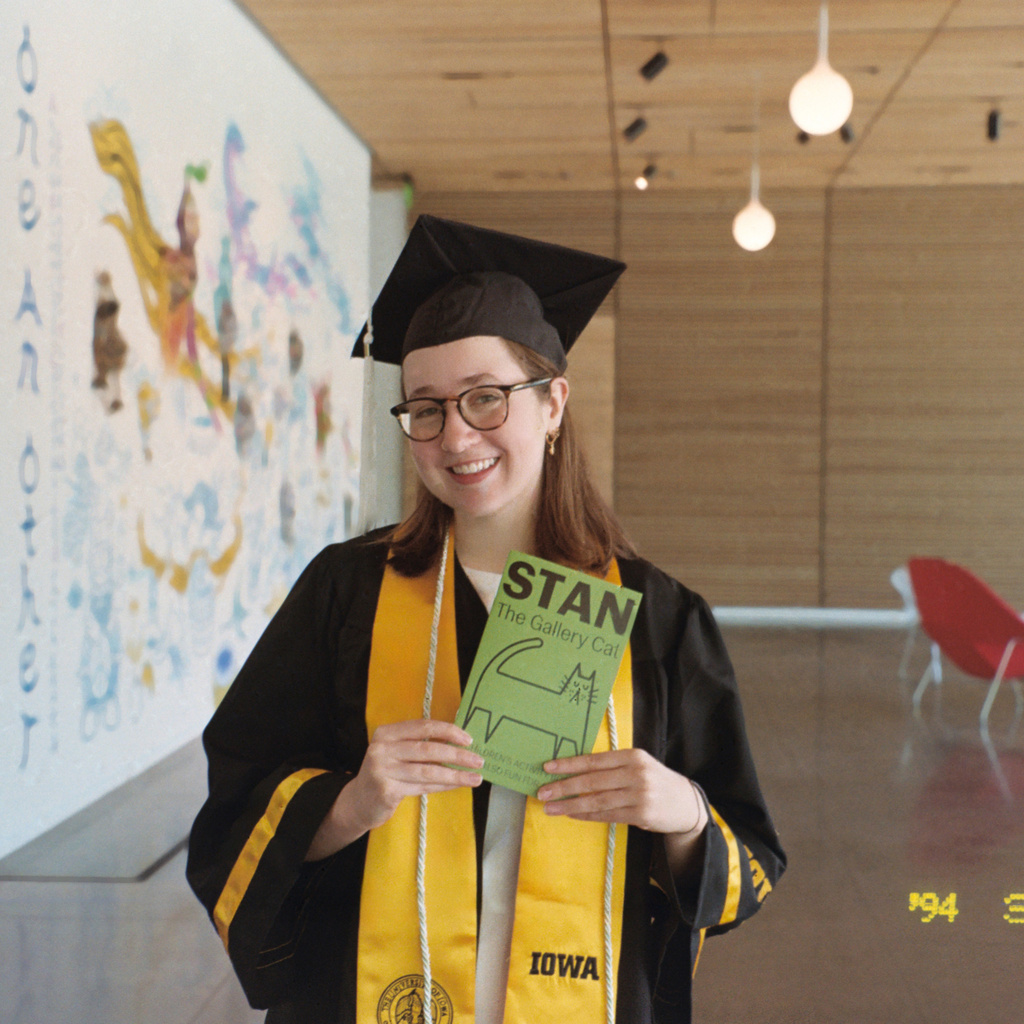 Museum Programs Intern Abbie McLaren, dressed in her graduation attire, poses with a Stan the Cat activity booklet, which she created in the spring of 2025, in the Stanley Museum of Art lobby.