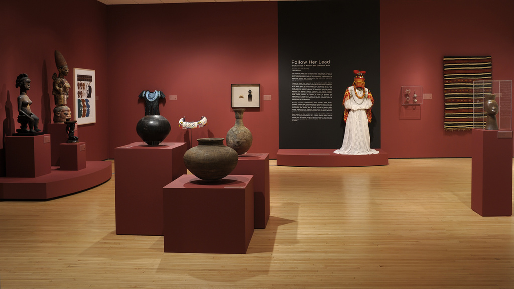 A museum gallery with dark red walls and light, wooden floor. There are large, African, ceramic pots in the foreground, and a mannequin with a white dress and jewelry in the background.